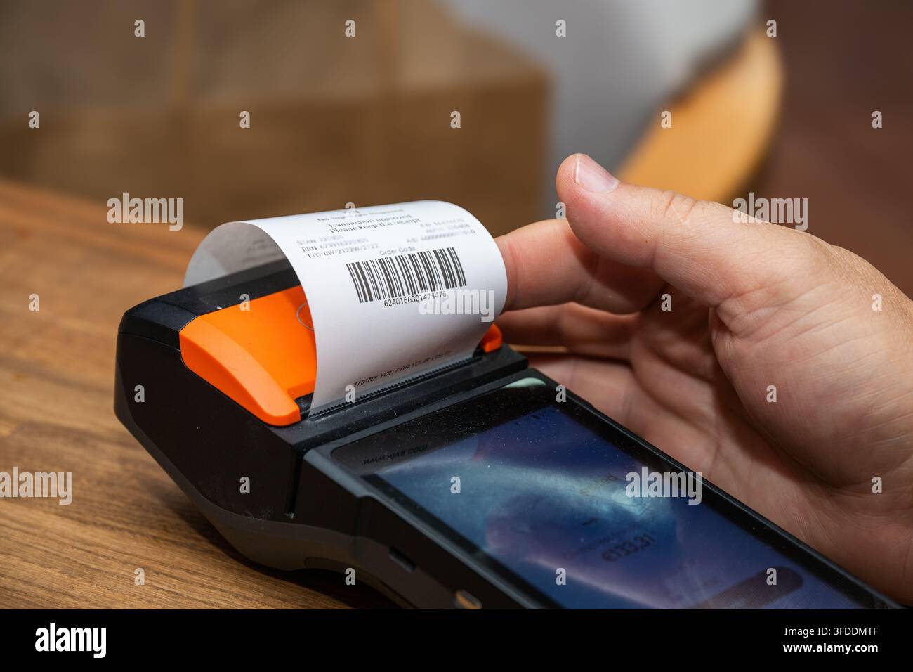 Una mano tiene una ricevuta stampata che fuoriesce da un terminale di pagamento POS moderno appoggiato su un bancone di legno. Foto Stock