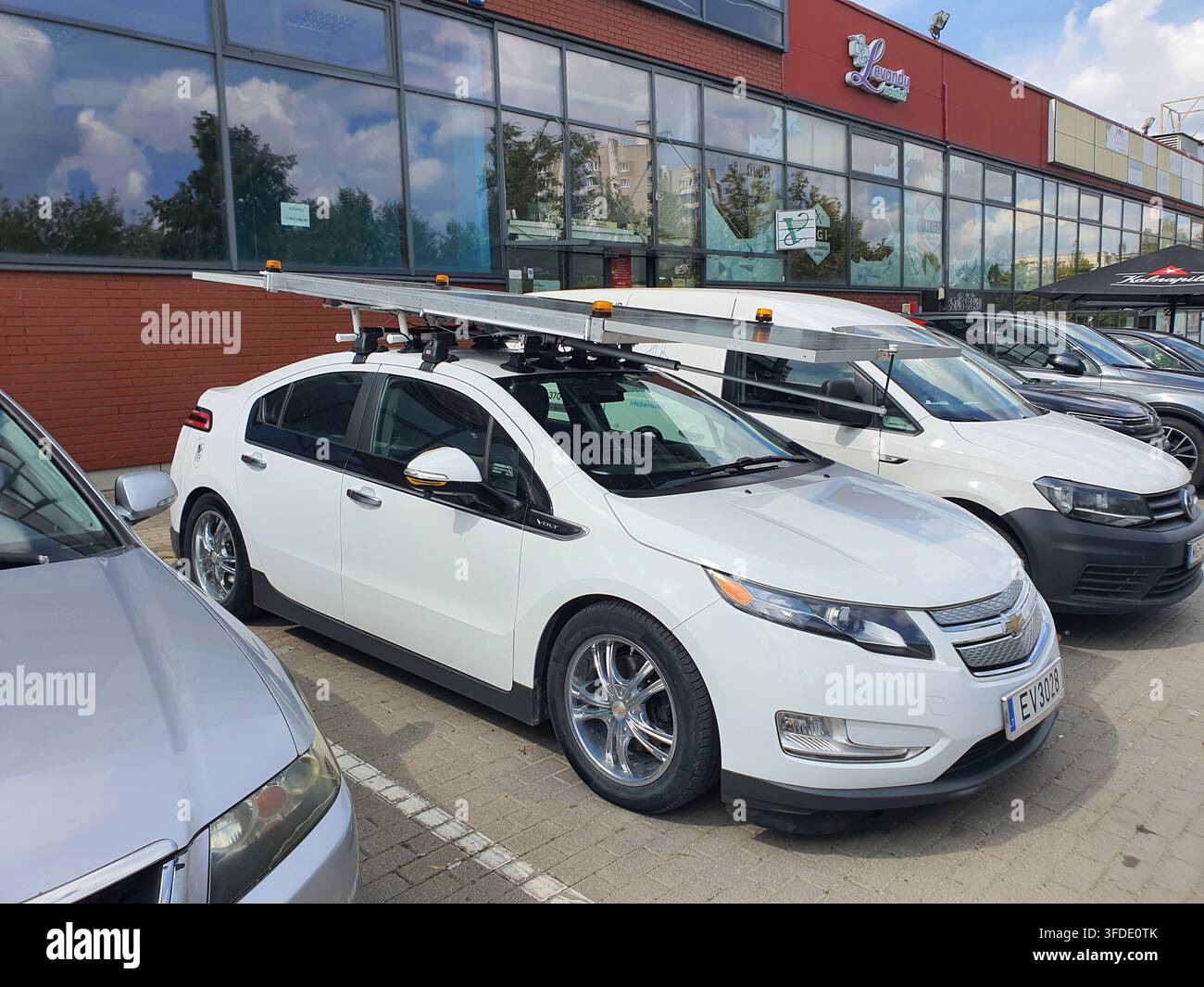 Un'auto ibrida plug-in bianca Chevrolet Volt dotata di portapacchi sul tetto che trasporta lunghe scale o travi metalliche è parcheggiata in un parcheggio lastricato. La Foto Stock