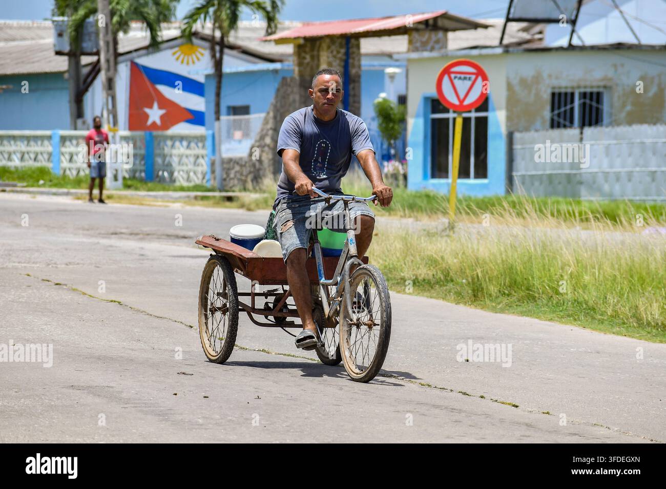 Cuba - Luglio 2026: Uomo in sella a un triciclo da carico modificato su una strada soleggiata con un murale della bandiera cubana sullo sfondo Foto Stock