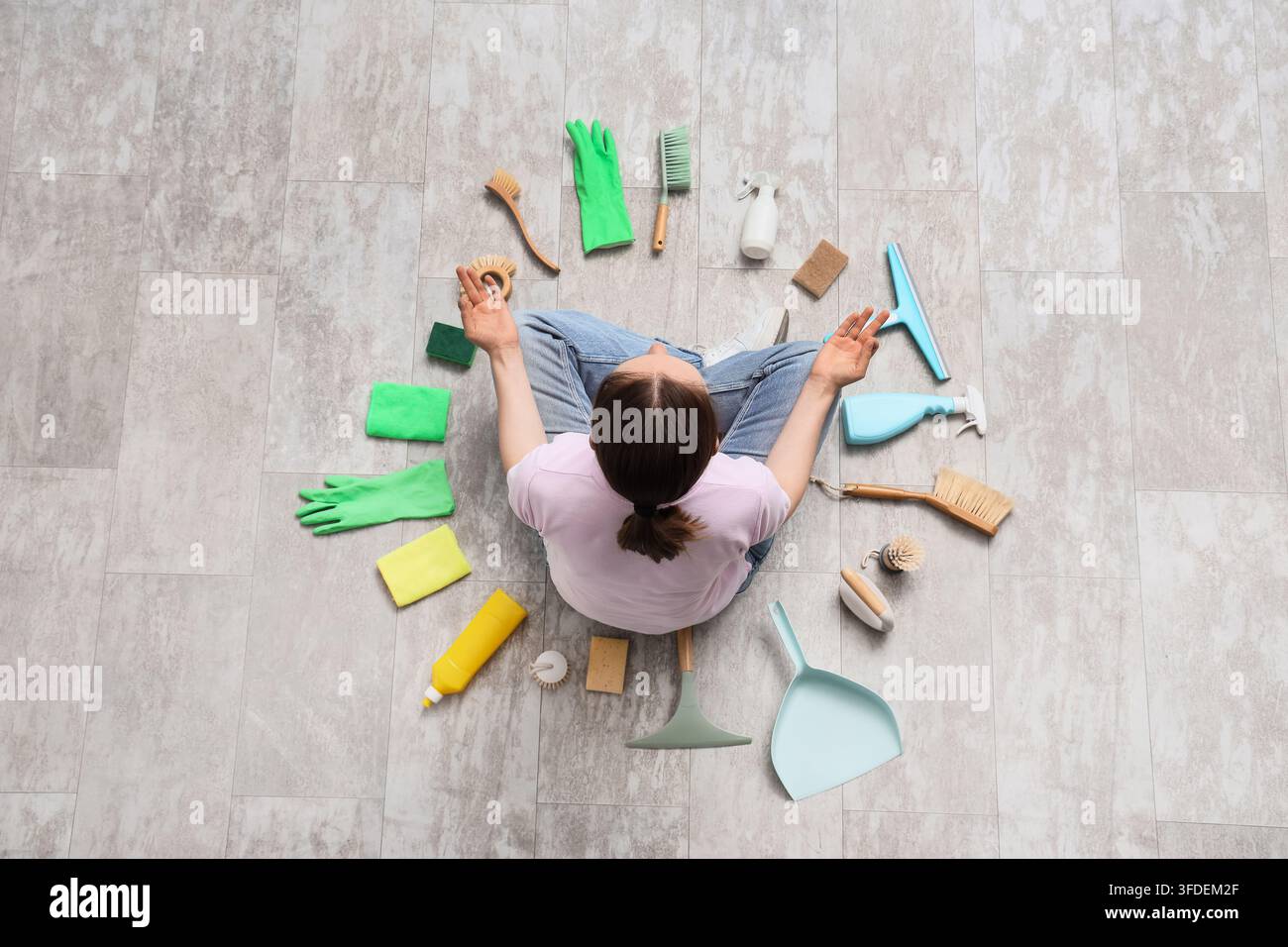Pulizia consapevole e sollievo dallo stress, donna seduta in posizione di meditazione circondata da faccende domestiche e prodotti per la pulizia su un pavimento piastrellato Foto Stock