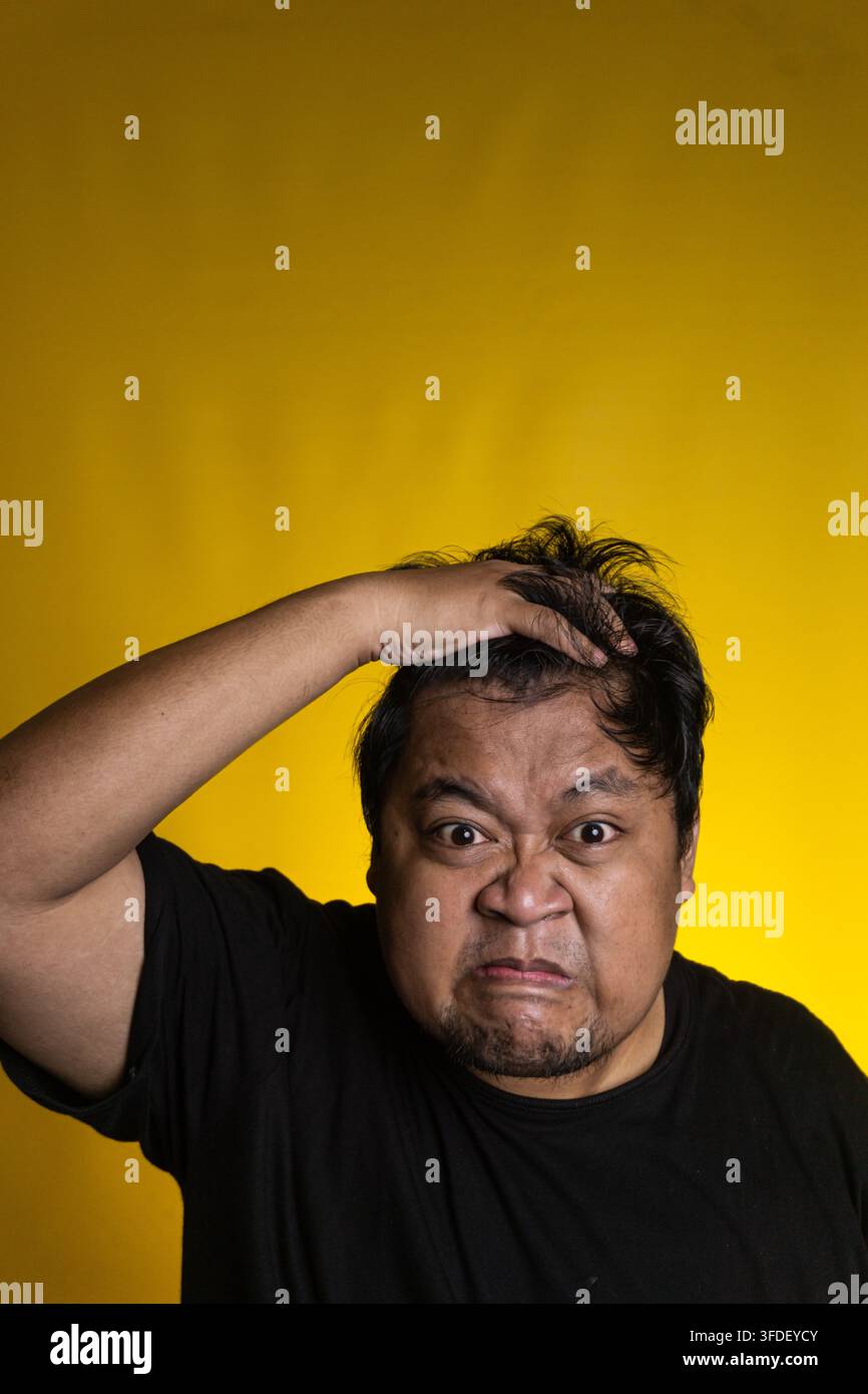 Un giovane uomo asiatico intensamente frustrato afferra aggressivamente i capelli con una mano mentre presenta un'espressione facciale ferocemente contratta contro un s Foto Stock