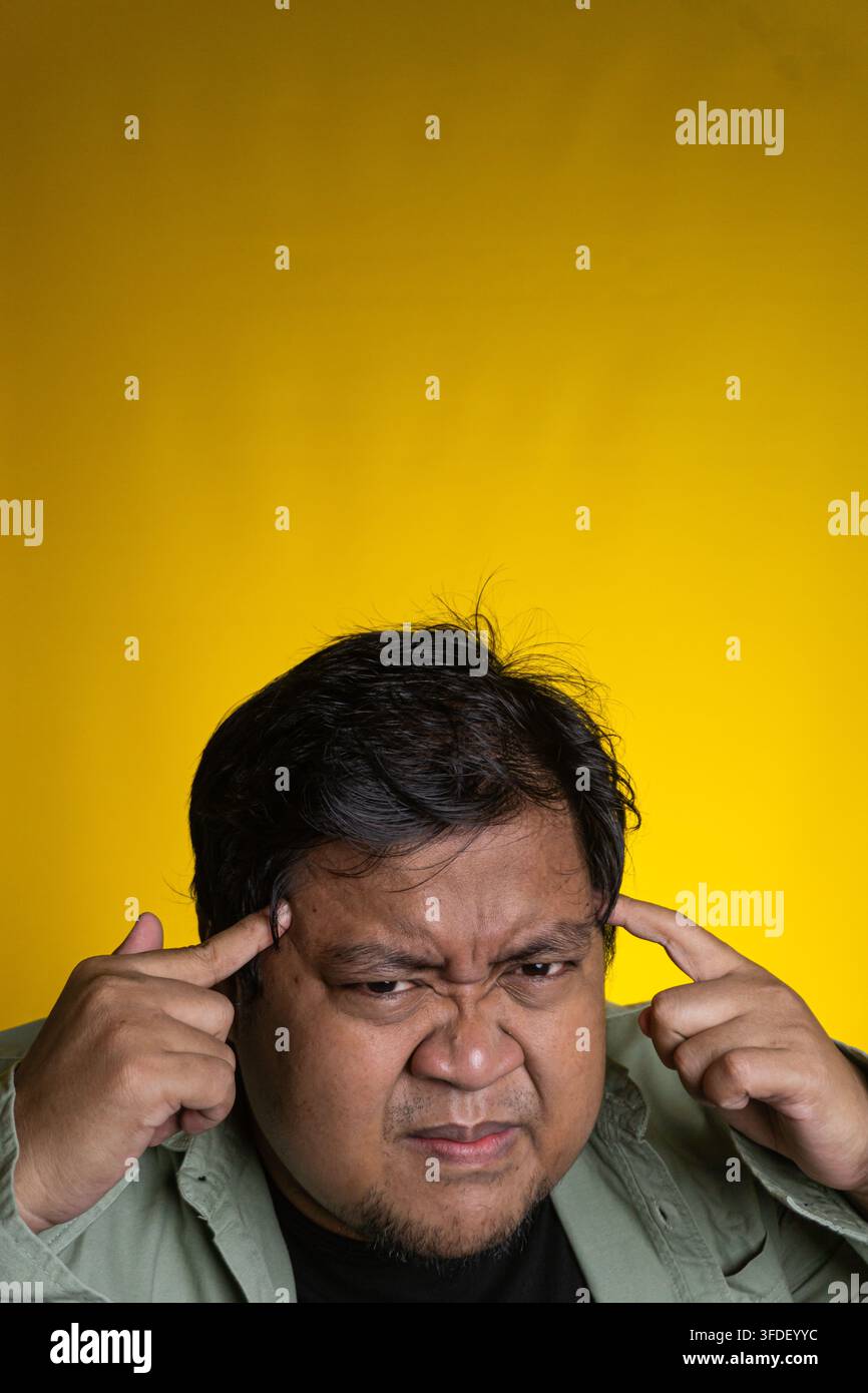 Un giovane uomo asiatico intenso preme entrambi gli indici contro le tempie mentre aggrotta profondamente le sopracciglia su uno sfondo giallo vivace. Lo stressato ind Foto Stock