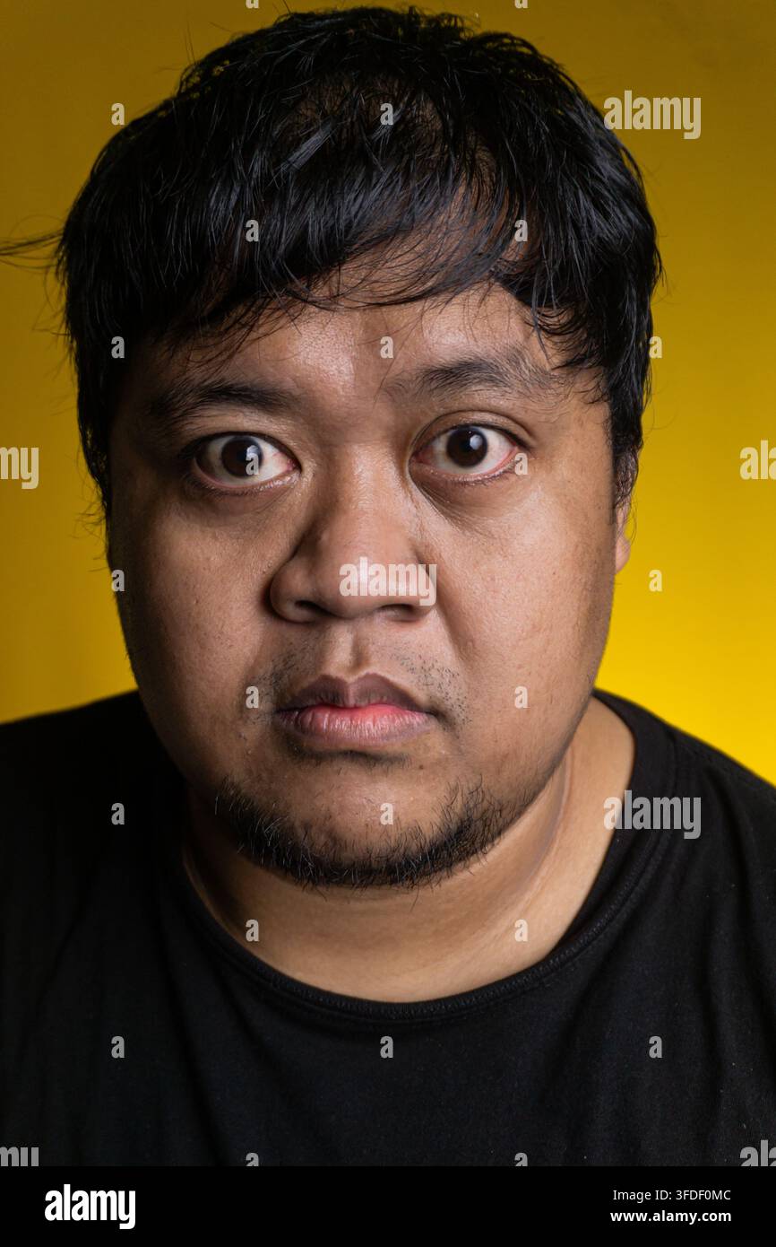 Un giovane uomo asiatico intenso fissa direttamente la fotocamera con gli occhi spalancati su uno sfondo giallo solido e vivace. L'individuo concentrato presenta Foto Stock