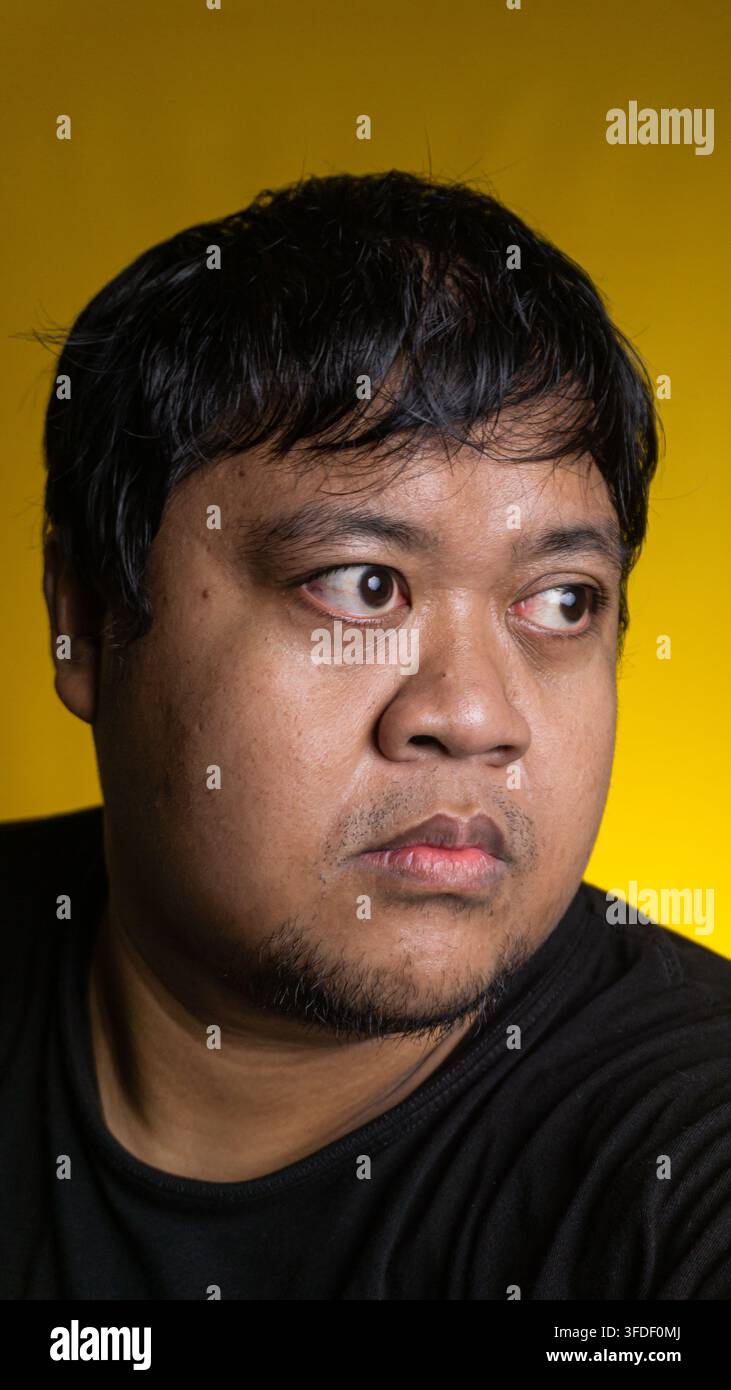 Un giovane uomo asiatico curioso guarda di lato con un'espressione seria su uno sfondo giallo solido e luminoso. L'individuo osservatore Foto Stock