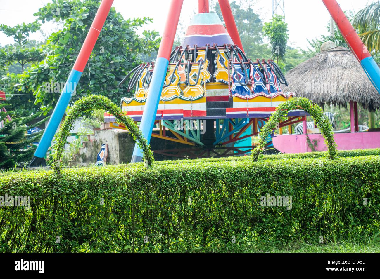 Vibrante attrazione da luna park in rotazione incorniciata da archi di siepi verdi ben curati in un parco soleggiato. Destinazione ricreativa familiare divertente con alberi tropicali. Foto Stock