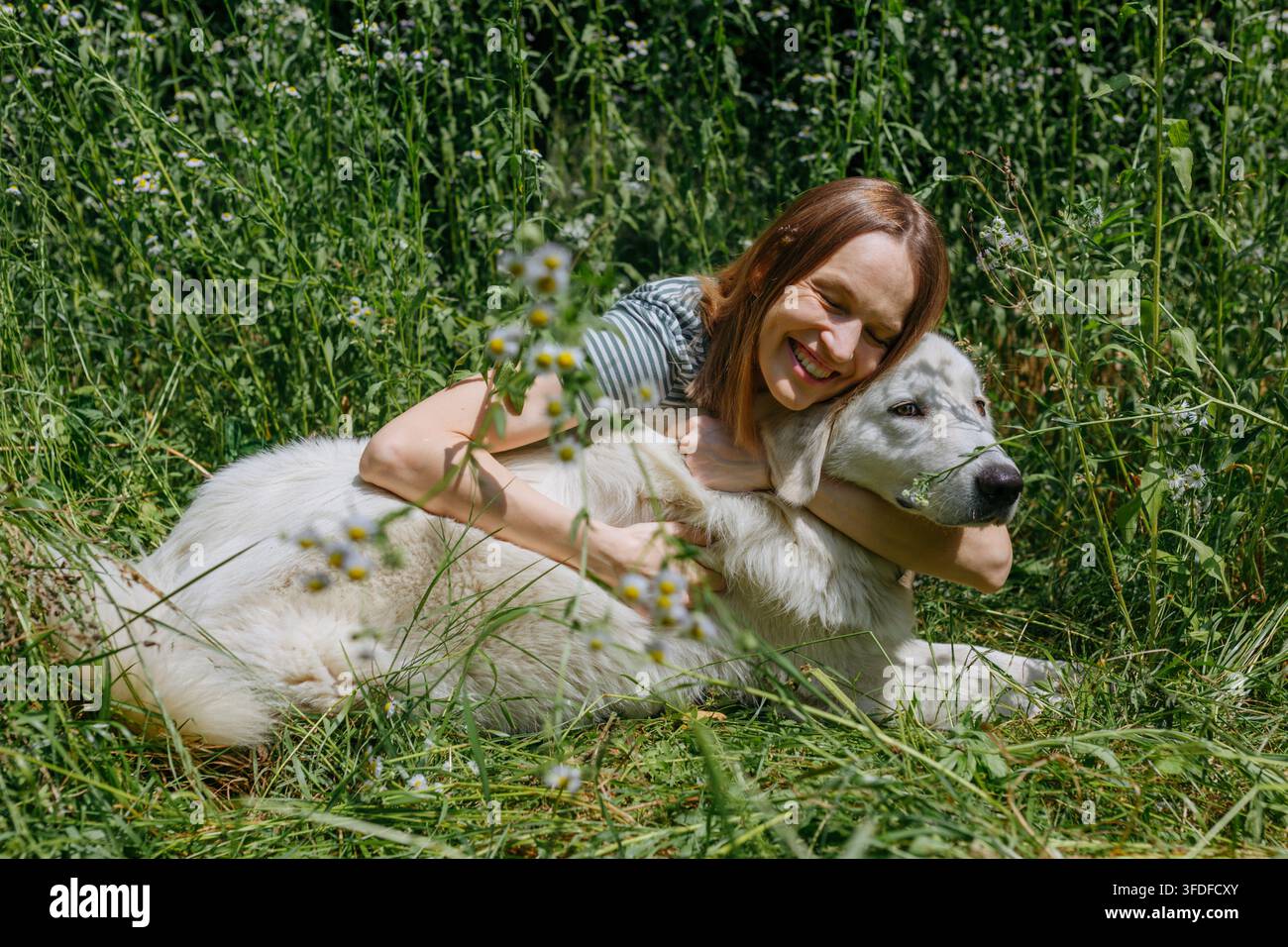 Donna gioiosa che abbraccia un cane da pastore Maremma in un prato di fiori selvatici Foto Stock