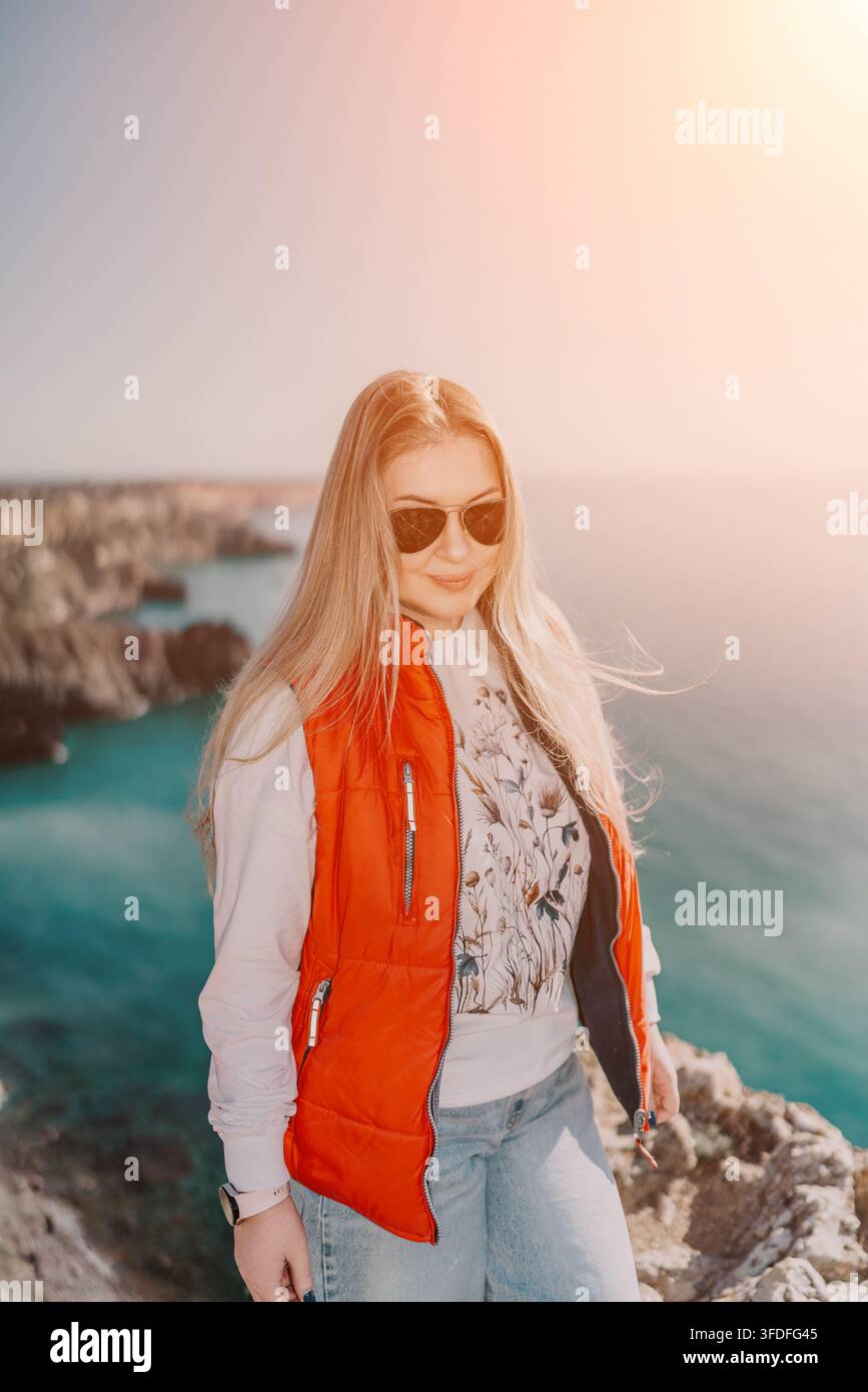 Donna bionda in un giubbotto rosso in piedi su una scogliera rocciosa che si affaccia sul mare durante l'ora dorata Foto Stock