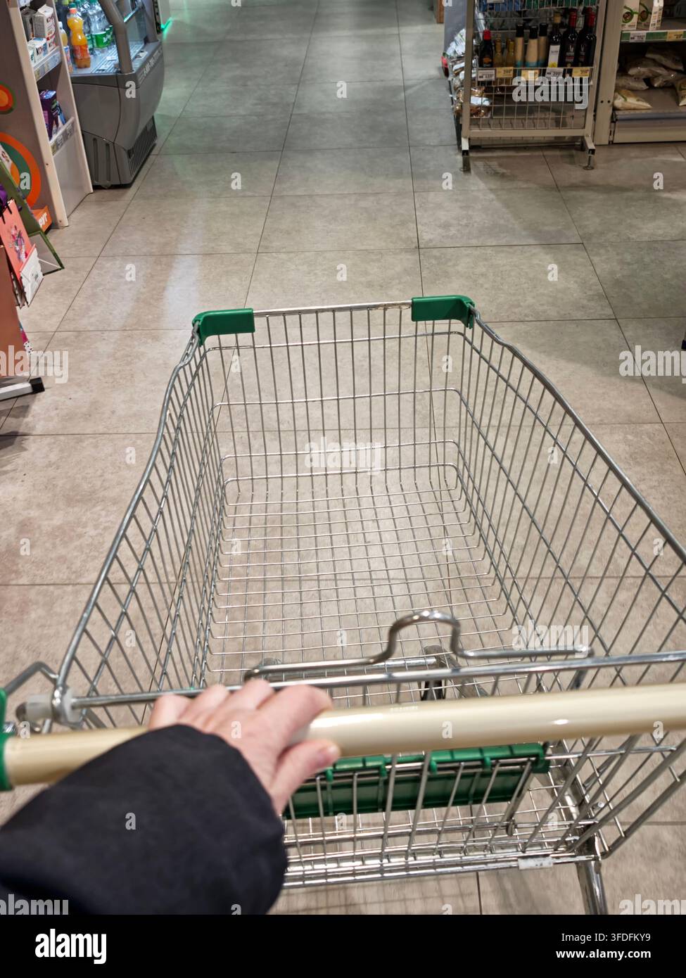 Kirov, Russia - 06 aprile 2026: Una persona tiene la maniglia di un carrello della spesa vuoto mentre cammina in un corridoio del supermercato cercando articoli da acquistare. Foto Stock