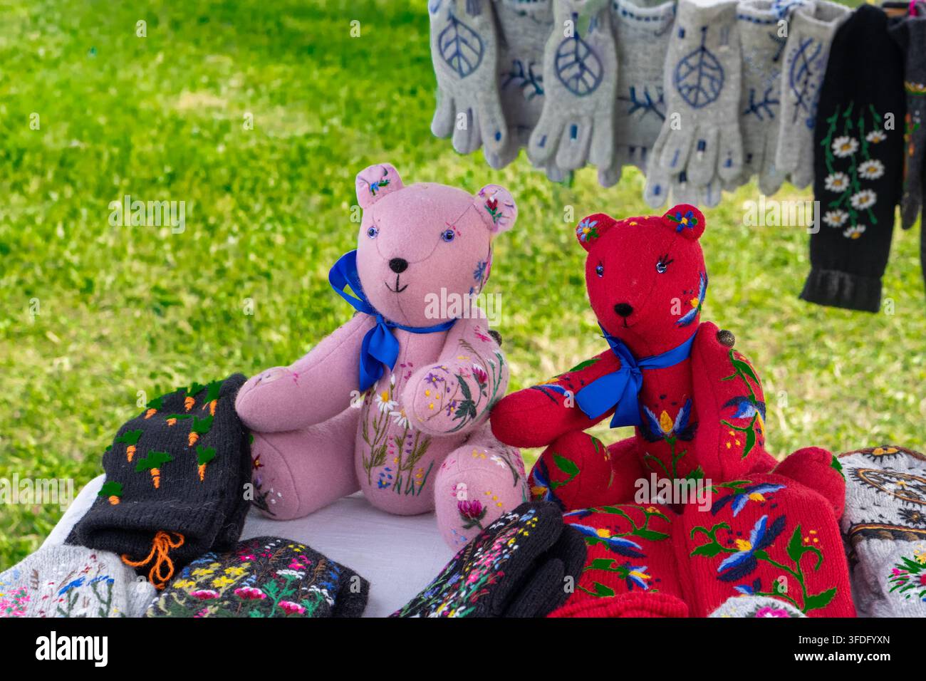 Orsetti di peluche rosa e rossi fatti a mano con ricami floreali esposti su un tavolo a una fiera artigianale all'aperto. Giocattoli morbidi in stoffa a forma di orso con fiocchi di nastro blu seduti Foto Stock