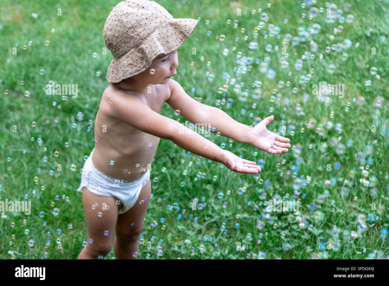 Un bambino con cappello da sole e pannolino è in piedi a piedi nudi sull'erba e allunga la mano verso numerose bolle di sapone fluttuanti. Adatto per articoli da gioco all'aperto, infanzia Foto Stock