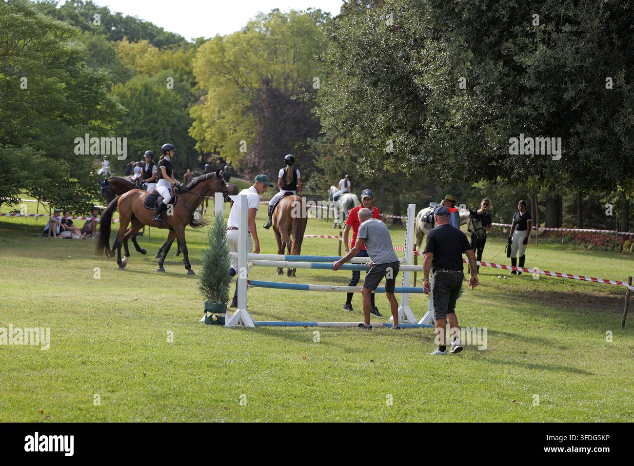 Equestri a cavallo aspettano in un'arena verde all'aperto mentre il personale regola un recinto di salto durante un evento di salto a ostacoli soleggiato. Bulgaria, Varna, 30 agosto 2026 Foto Stock