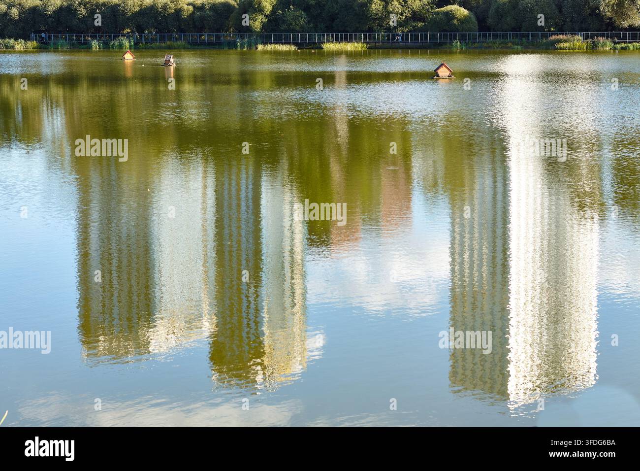 Gli edifici moderni si riflettono nella calma acqua del lago, circondato da una vegetazione rigogliosa, mentre delle casette per anatre in legno galleggiano in superficie. Foto Stock