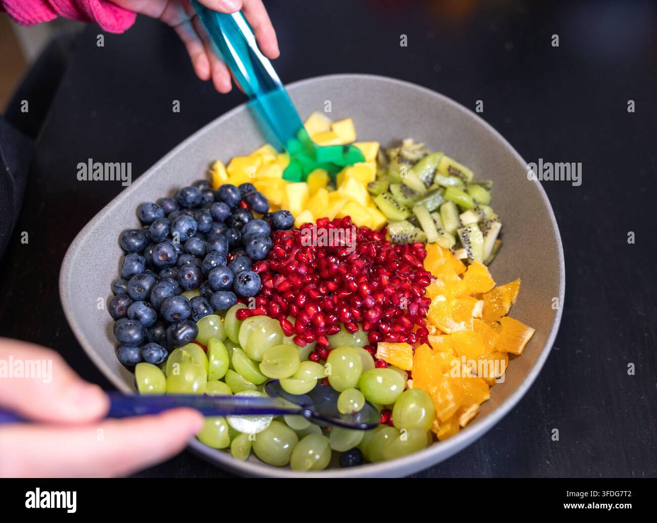 Donna mescola kiwi, arance, ananas, mirtilli, uva e semi di melograno in una ciotola per fare un'insalata di frutta. Donna mescola kiwi, arance, ananas Foto Stock