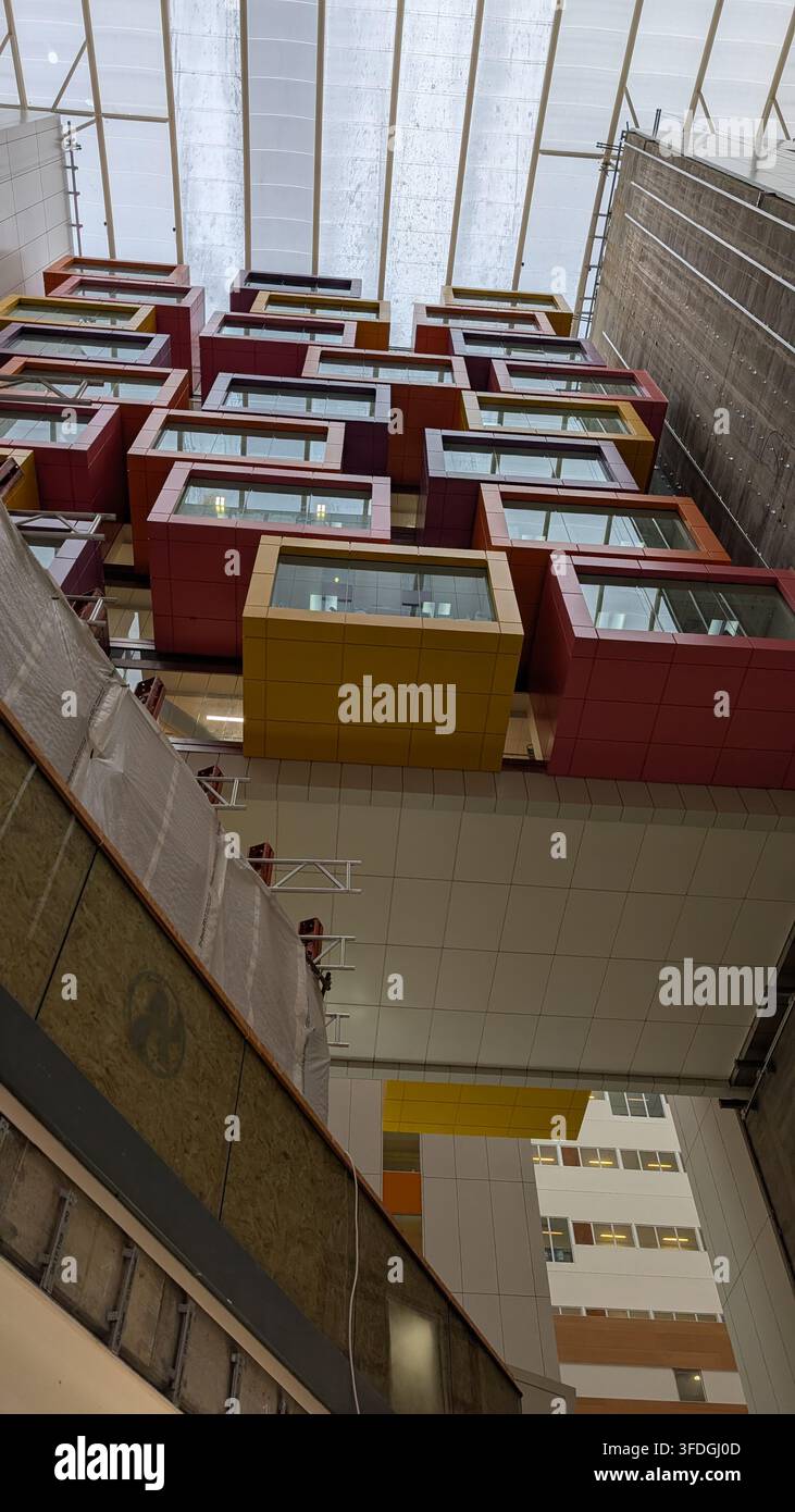 Una veduta a bassa angolazione mette in risalto un vasto atrio interno moderno con un soffitto a lucernario in vetro e file di vivaci pod di ufficio geometrici e colorati impilati Foto Stock