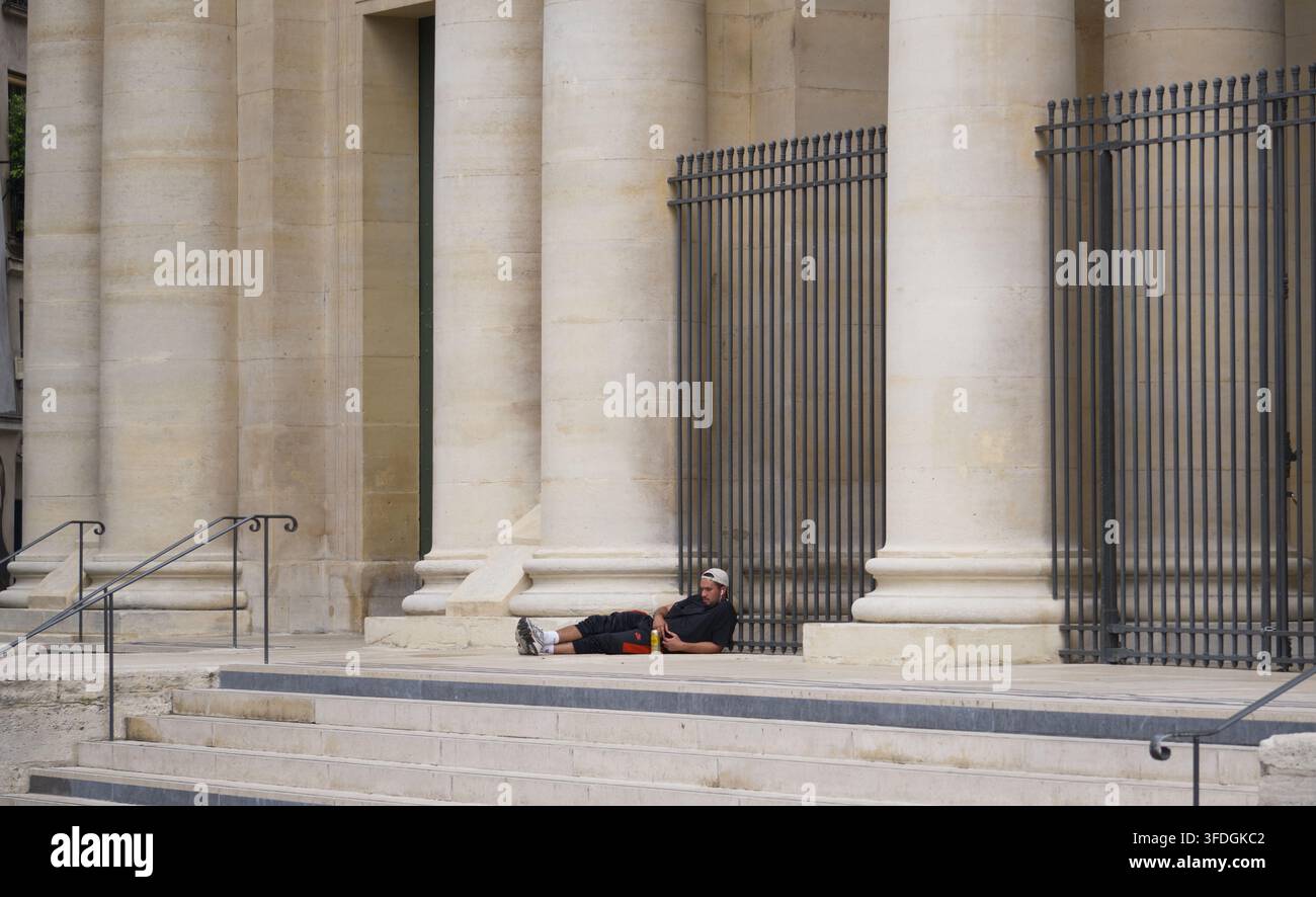 Un uomo sdraiato e riposante sui gradini all'esterno di un edificio classico in pietra con grandi colonne e cancelli di ferro a Parigi, Francia. Foto Stock
