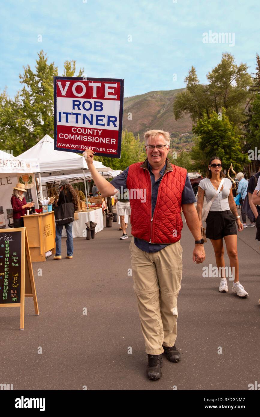 Uomo con giacca rossa porta un cartello in un mercato all'aperto che recita "Vota Rob Ittner per il Commissario della Contea di Pitkin, Aspen, Colorado." Foto Stock