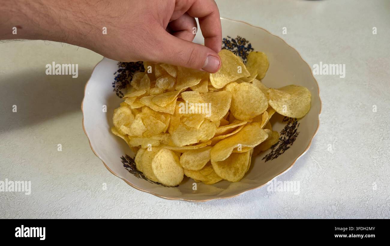 Mano che raggiunge una patatina fritta croccante in una ciotola, che illustra lo spuntino casual, il finger food e il mangiare indulgente. Foto Stock