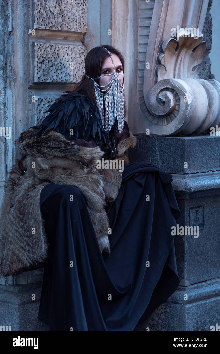 Personaggio gotico seduto su un piedistallo di pietra con mantello di pelliccia e piume, scatto narrativo cinematografico. Immagine di eroina dark fantasy per un poster o copertina di libro Foto Stock