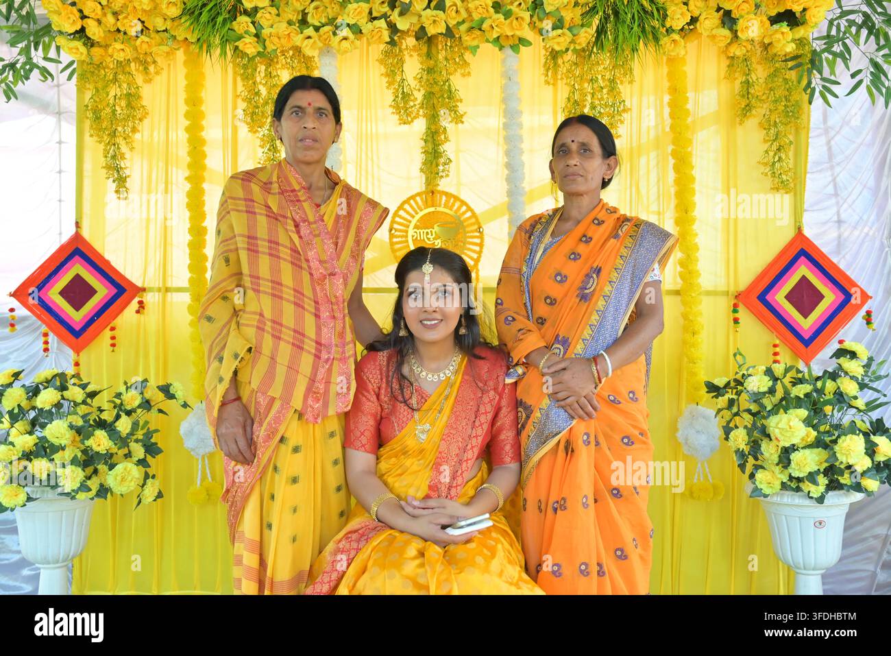 Sposa indiana in saree giallo seduta tra due donne anziane durante la cerimonia Haldi a una funzione nuziale bengalese con decorazioni di fiori di calendule gialli Foto Stock