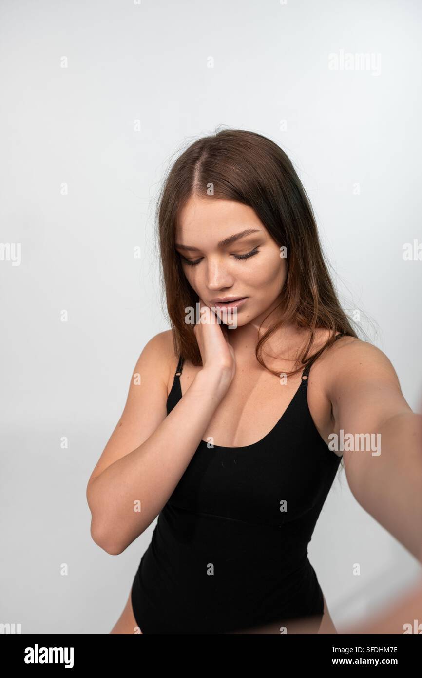 Giovane donna che si scatta un selfie, si tocca il collo, mostra bellezza naturale e amore per sé stessa su sfondo bianco Foto Stock