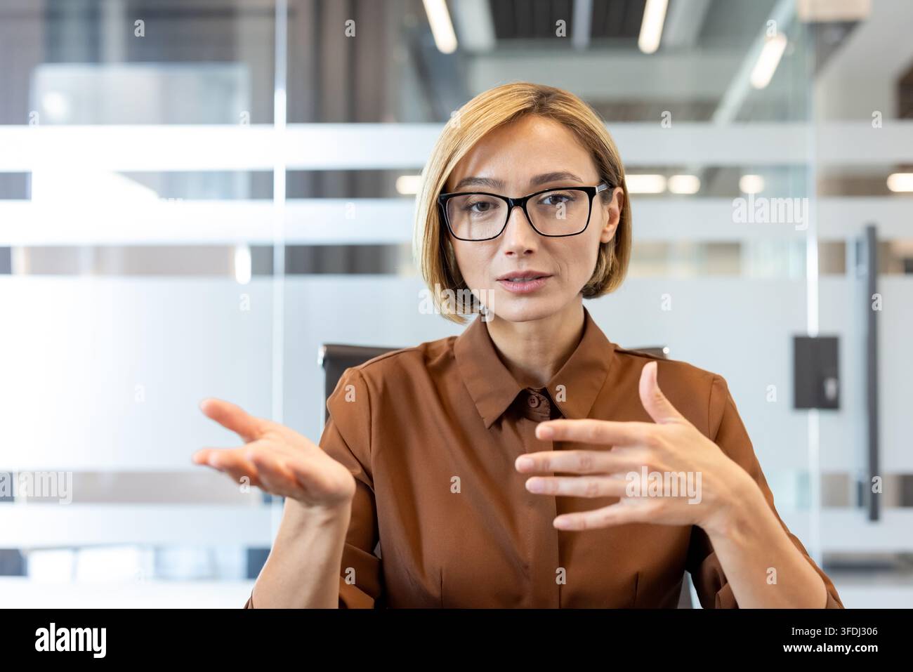 Donna d'affari sicura di sé con occhiali che comunica e gesticola attivamente durante una conferenza virtuale o una riunione online, mostrando professionalità e leadership in un ambiente di ufficio moderno Foto Stock