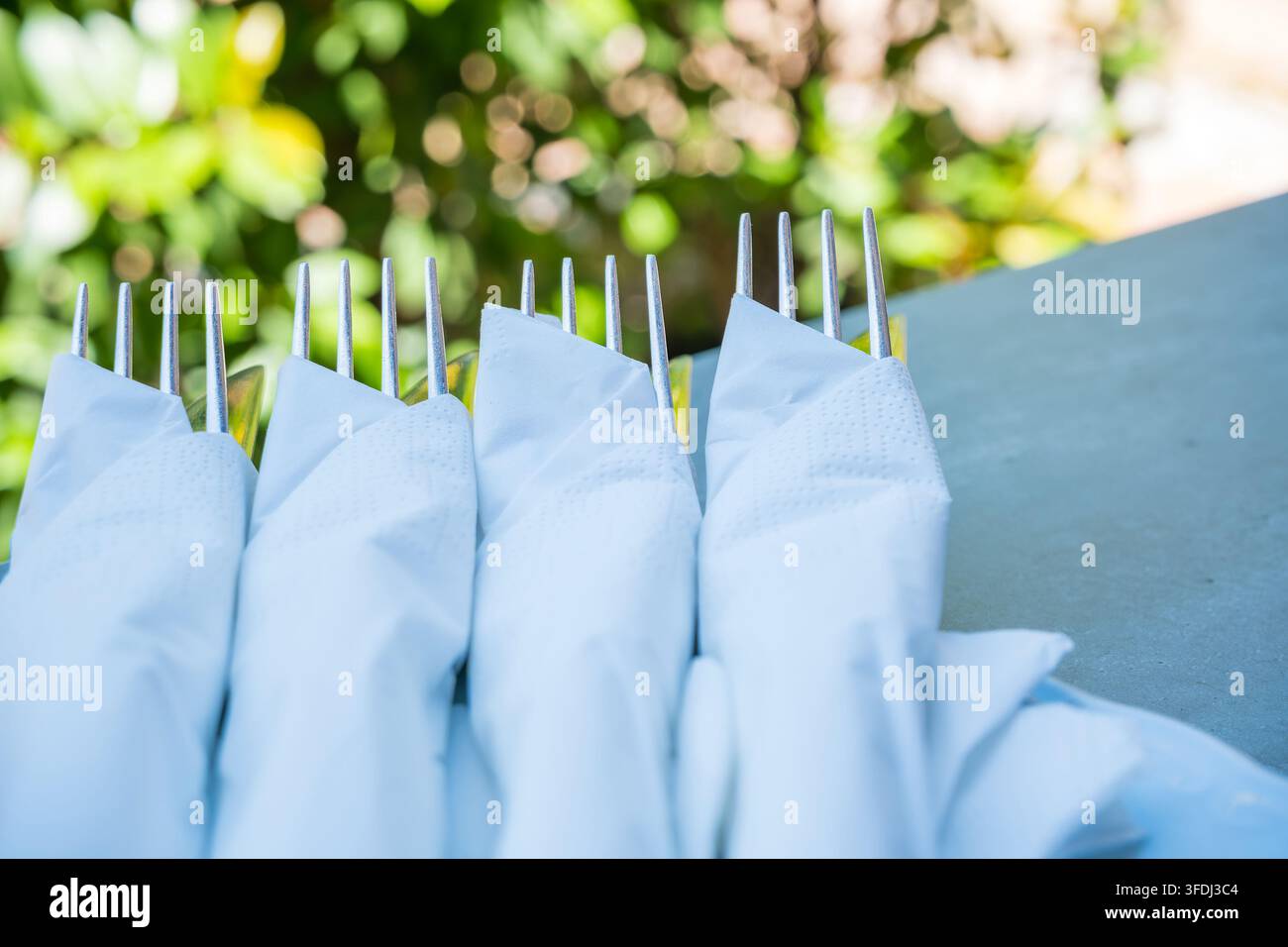 Tovaglioli di stoffa bianchi avvolti intorno a forchette d'argento in fila su un tavolo esterno blu, con una morbida sfondo di fogliame verde illuminato dal sole Foto Stock