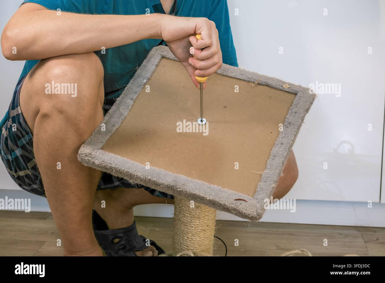 Persona che utilizza un cacciavite giallo per fissare la base tappezzata di un tiragraffi avvolto in sisal al chiuso su un pavimento di legno Foto Stock
