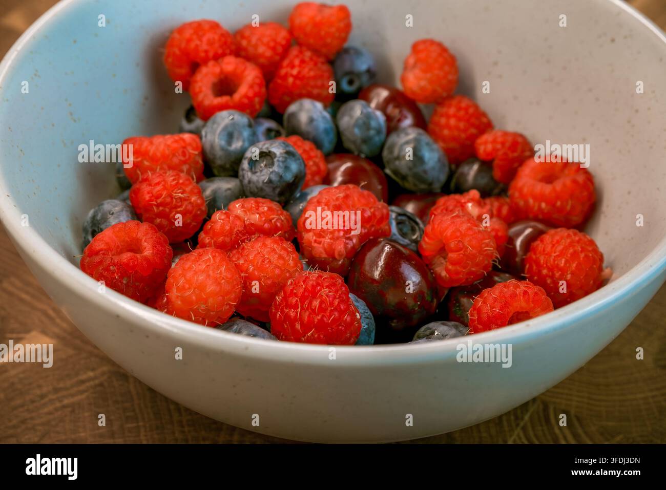 Prima vista di lamponi, mirtilli e ciliegie disposti in una ciotola blu e bianca maculata su un tavolo di legno con frutta mista colorata Foto Stock