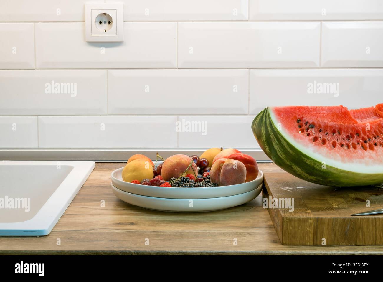 Fetta grande di anguria e ciotola di pesche, nettarine, lamponi, mirtilli e ciliegie su un piano di lavoro della cucina in legno accanto a un peccato Foto Stock