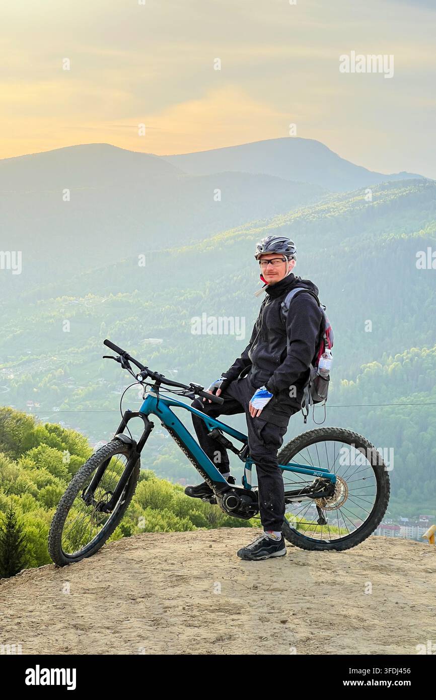Uomo seduto su una mountain bike elettrica sulla vetta di una collina, ciclista che posa con una valle montana scenografica e un cielo al tramonto, concetto di avventura all'aperto Foto Stock
