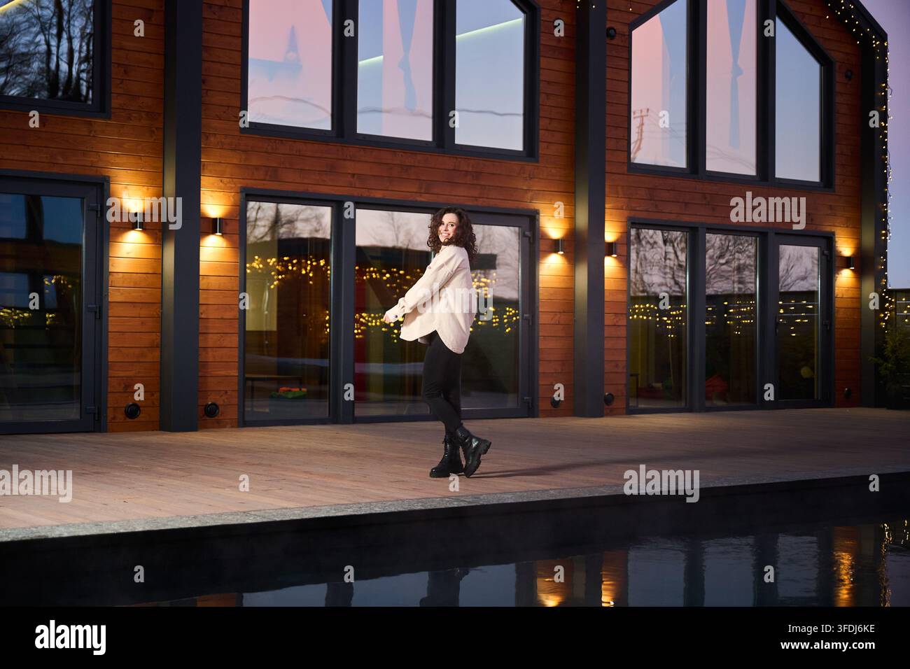 Donna sorridente che passeggia accanto a una moderna villa in legno con piscina e luci festive, ideale per temi di viaggi di lusso, immobili di fascia alta e uno stile di vita ambizioso Foto Stock