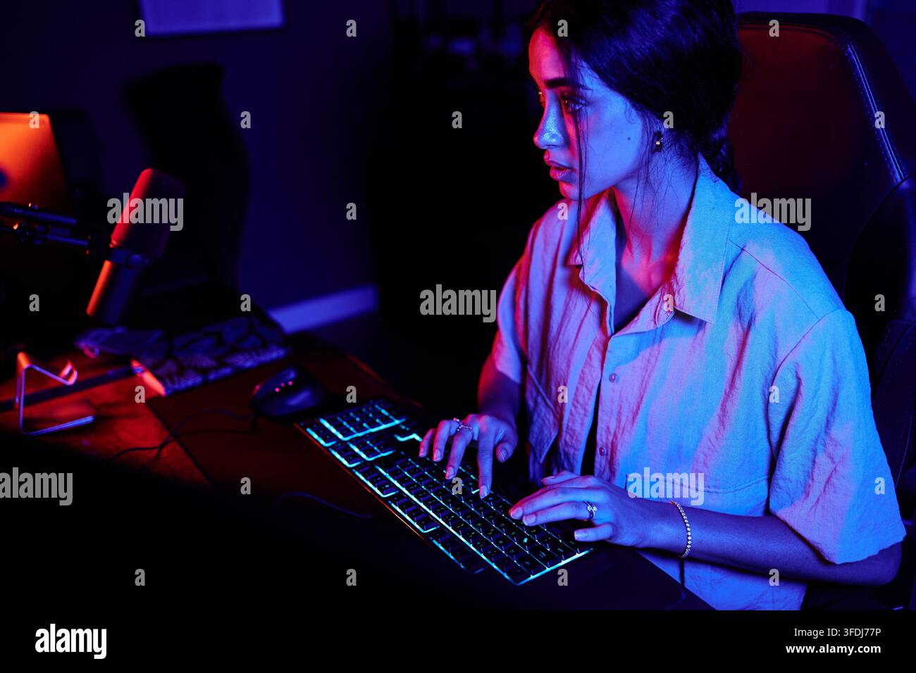 Donna che utilizza un setup da gaming con una tastiera retroilluminata in una stanza buia, illuminata da luci neon rosse e blu Foto Stock