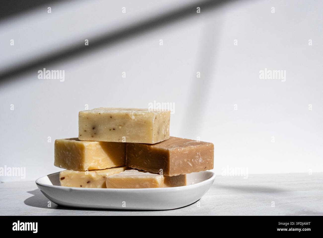 Pila di saponi artigianali fatti a mano disposti su un piatto di ceramica bianca con una luce naturale soffusa. Le barre rustiche presentano toni neutri caldi e naturali va Foto Stock
