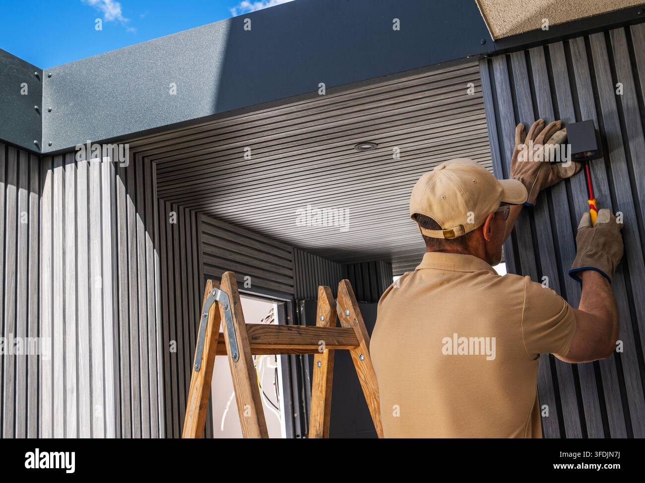 Un operaio tiene una lampada da parete contro il rivestimento esterno e stringe una vite sotto di essa. Una scala di legno è posizionata accanto all'ingresso coperto. Foto Stock
