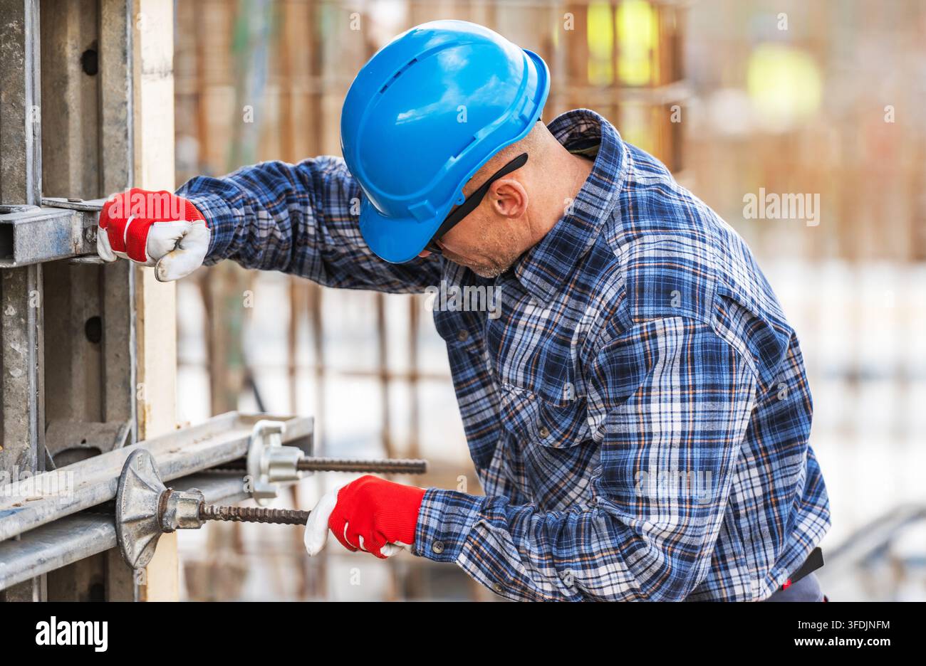 Un operaio con casco e guanti regola una morsetta metallica su una gettatura verticale in cemento. Dietro di lui appaiono reti di ferro e ulteriori elementi strutturali. Foto Stock
