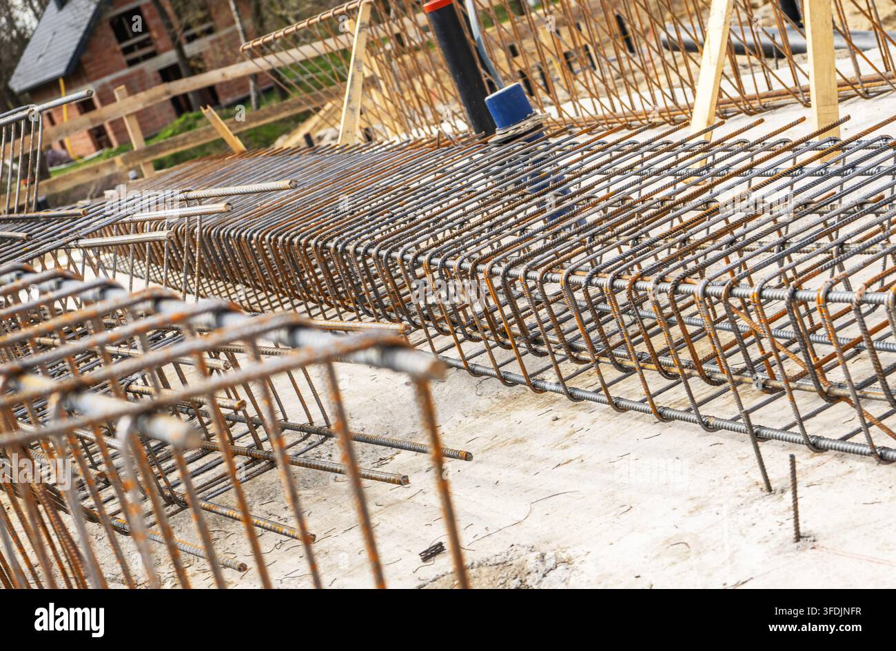 Righe ripetute di barre d'acciaio piegate si estendono su una lastra di cemento in un cantiere edile. Casseri in legno e strutture portanti riempiono lo sfondo. Foto Stock