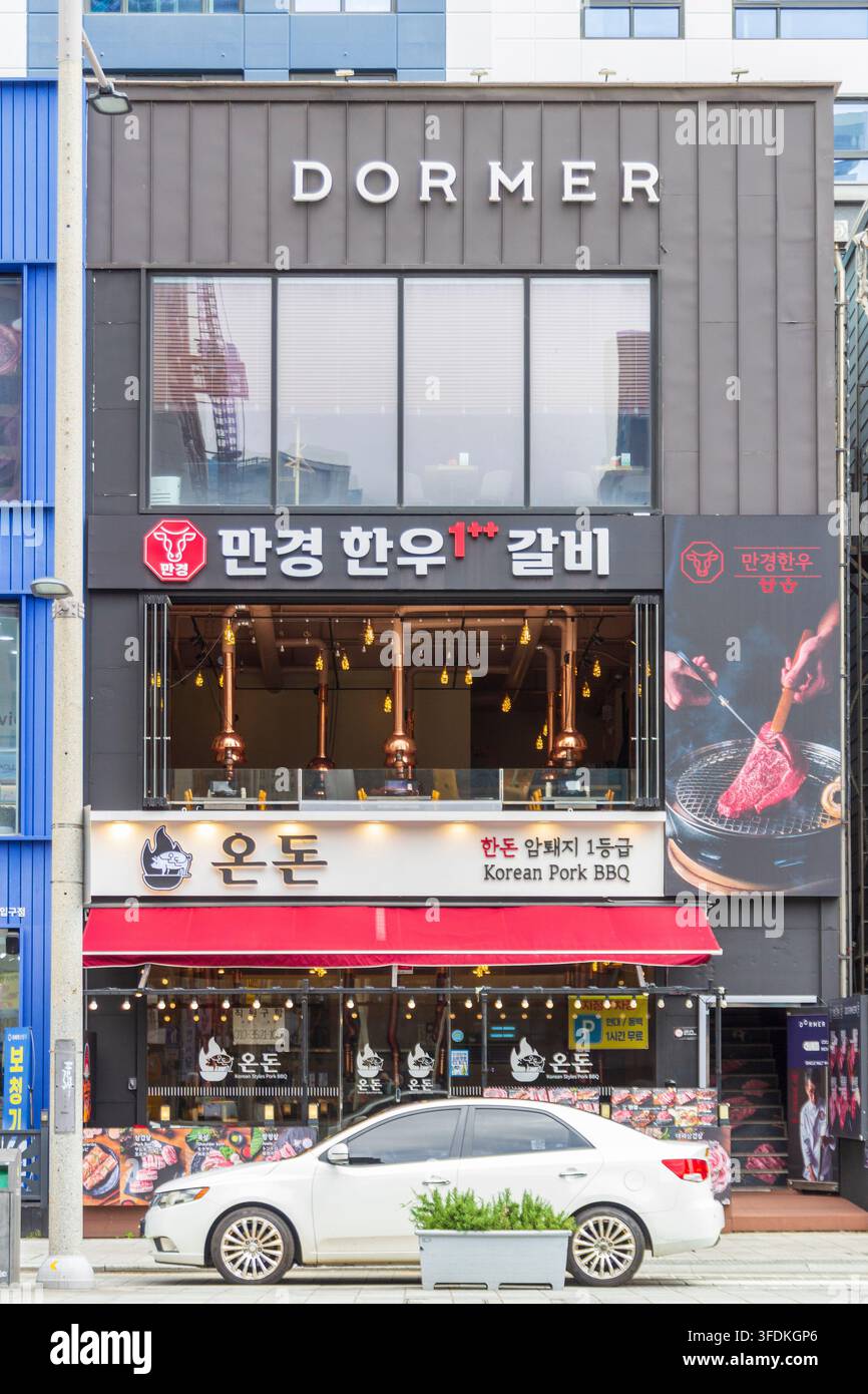 La facciata di un ristorante coreano di barbecue di maiale presenta cartelli in Hangul, una tenda rossa, grandi finestre, cappe estrattive per grill e una berlina bianca parcheggiata all'esterno. Foto Stock