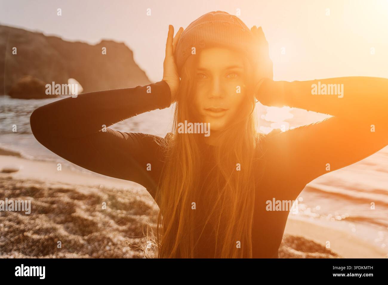 Donna, spiaggia, ritratto di surfista con berretto in un tramonto dorato con bagliore del sole su una riva rocciosa Foto Stock