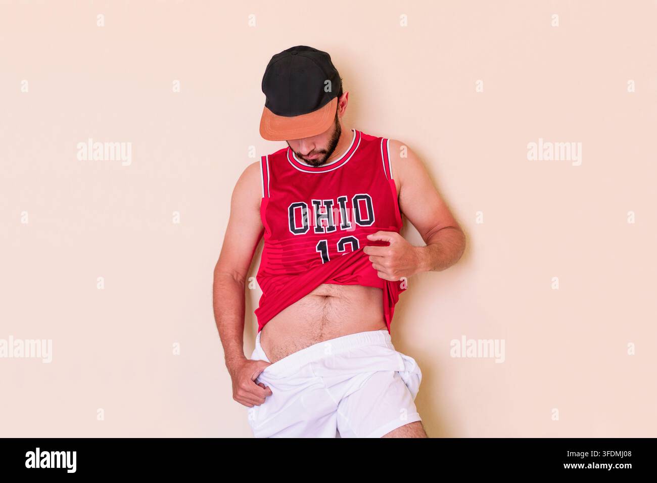 Giovane uomo atletico che indossa una canottiera rossa della squadra e un berretto nero, solleva la maglietta e abbassa i pantaloncini. Un tifoso sportivo maschio in pantaloncini bianchi; le sue gambe Foto Stock