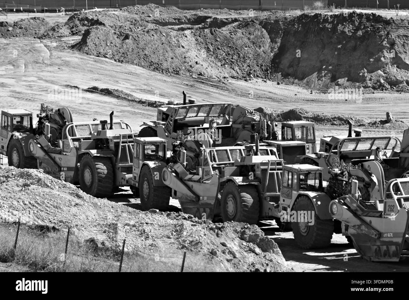 Linee di macchinari gialli su un cantiere polveroso. Le attrezzature pesanti riposano inattive sotto il sole luminoso. Cumuli di terra si alzano dietro i macchinari. Nessun lavoratore visibile Foto Stock