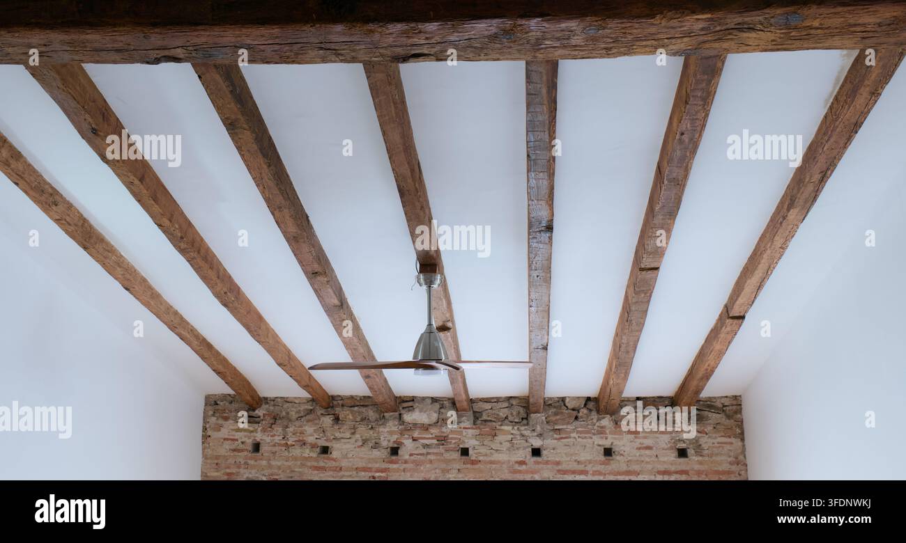 Una vista simmetrica suggestiva cattura un moderno ventilatore da soffitto sospeso sotto un soffitto bianco immacolato, interrotto da una serie di fissaggi in legno scuro e rustico Foto Stock