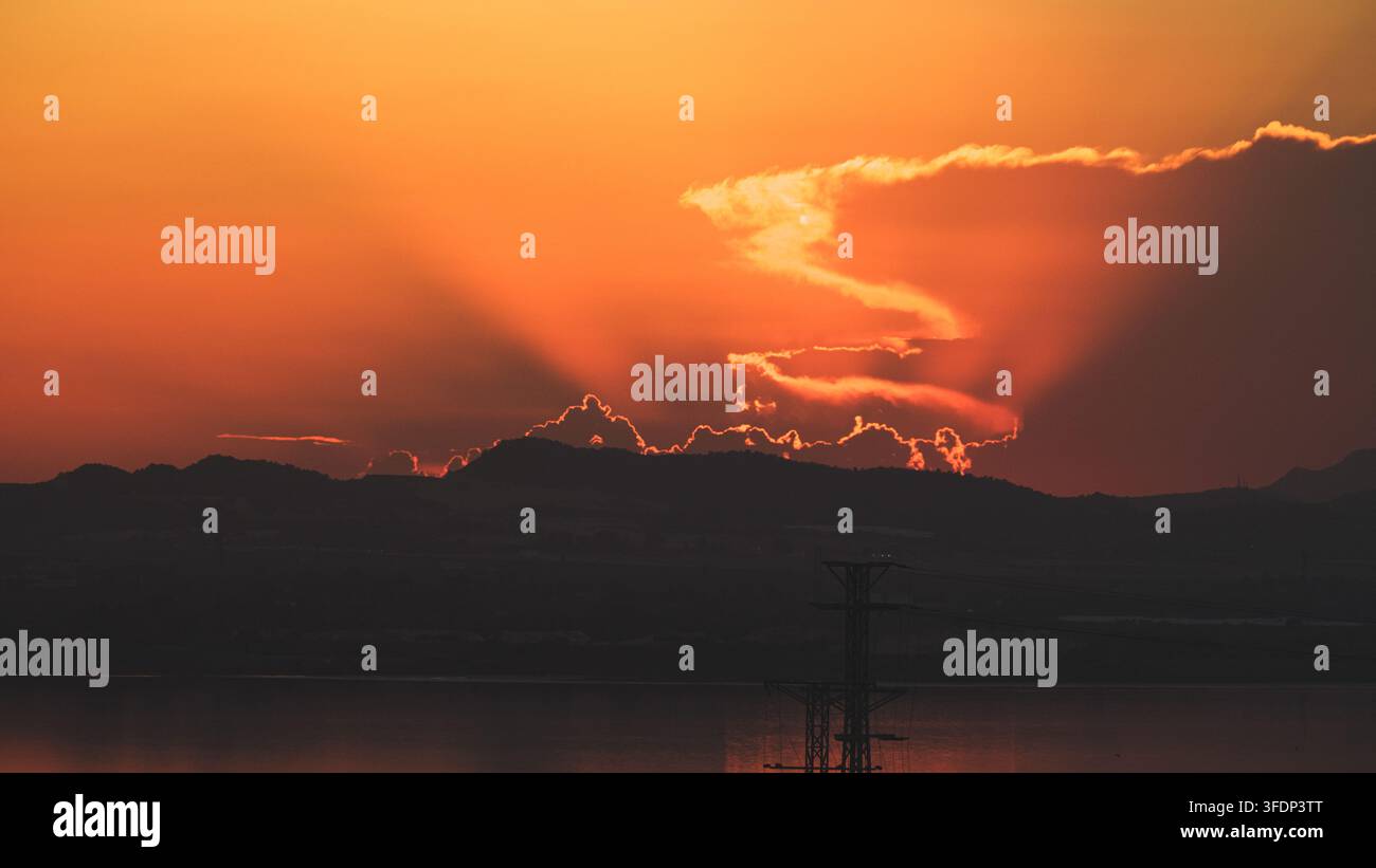 Un tramonto drammatico all'ora dorata proietta una luce arancione vibrante su una cresta montuosa scura e su un pylon elettrico prominente, creando un'atmosfera industriale serena Foto Stock