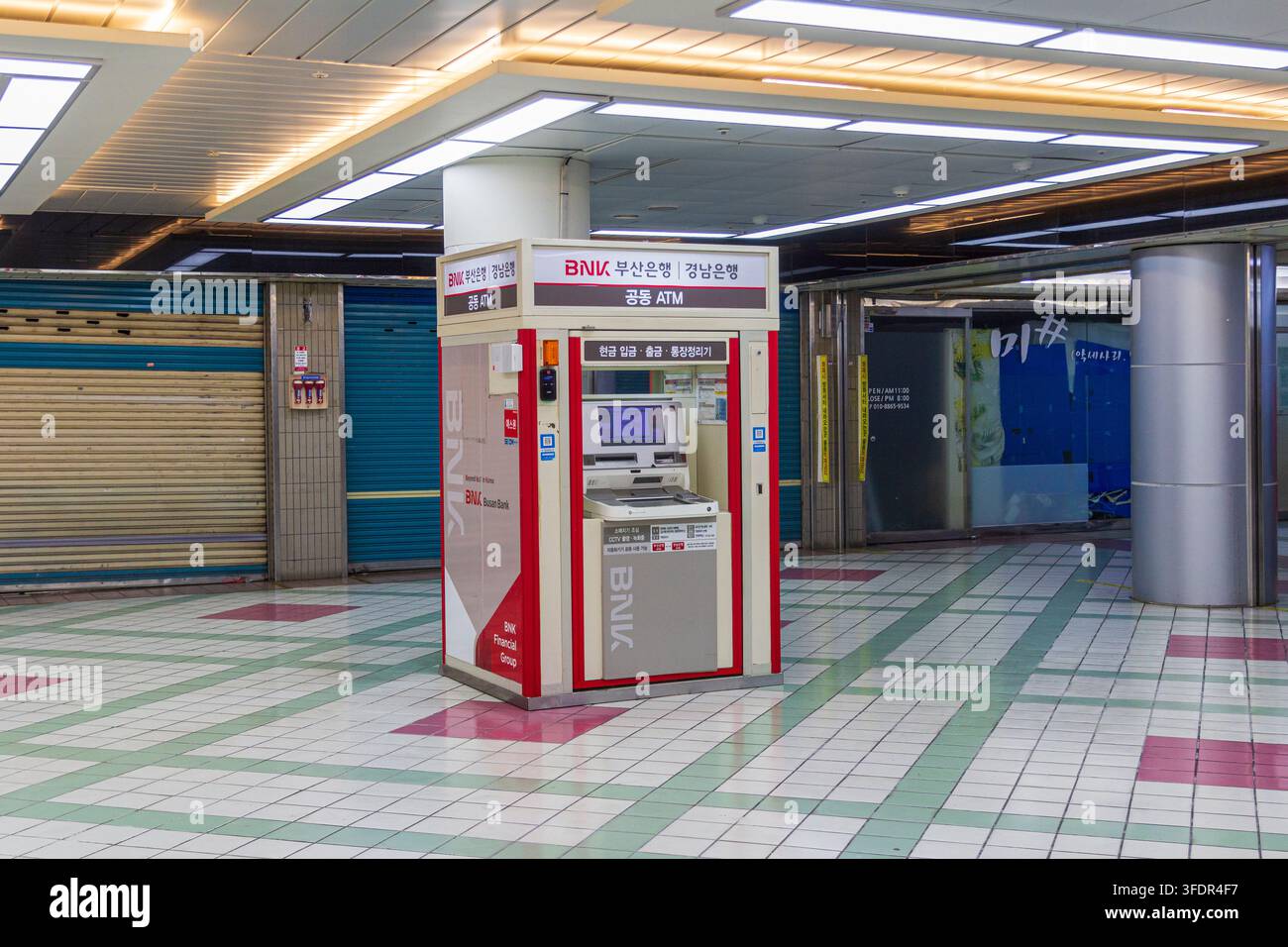 I chioschi bancomat di marca si trovano accanto a saracinesche metalliche chiuse sotto luci geometriche al soffitto in un centro commerciale coperto vuoto. Foto Stock