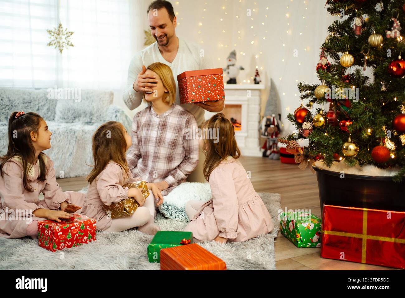 Padre che sorprende la madre con un regalo di Natale mentre le figlie guardano Foto Stock