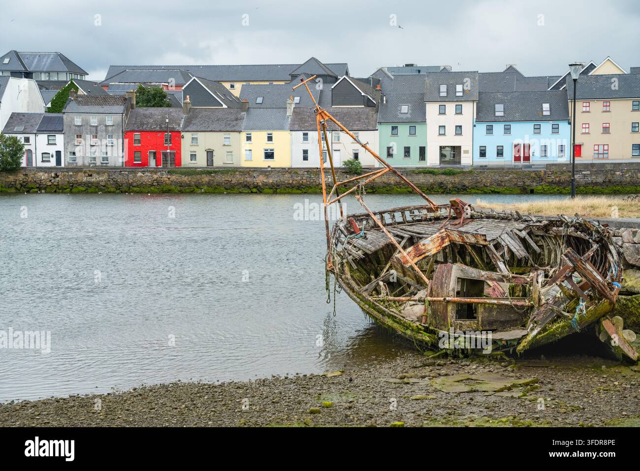 Una nave logorata accanto alla città vivace contrappone una barca di legno in decadenza alla bassa marea con il colorato lungomare di Long Walk a Galway per un ricordo marittimo Foto Stock