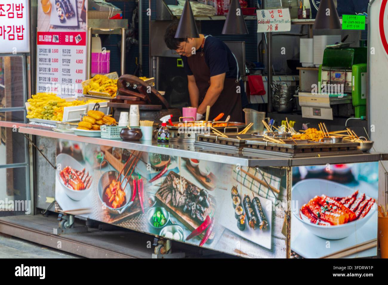 Due venditori preparano e servono vari cibi fritti e spiedini dietro il bancone di una bancarella di mercato coreana. Foto Stock