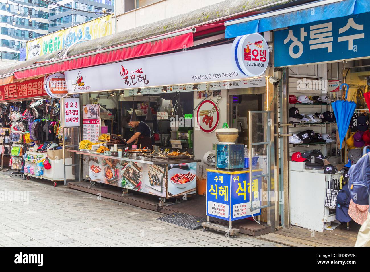Un venditore lavora dietro un banco di cibo che espone snack fritti e spiedini sotto cartelli del menu in lingua coreana in un mercato al dettaglio coperto. Foto Stock