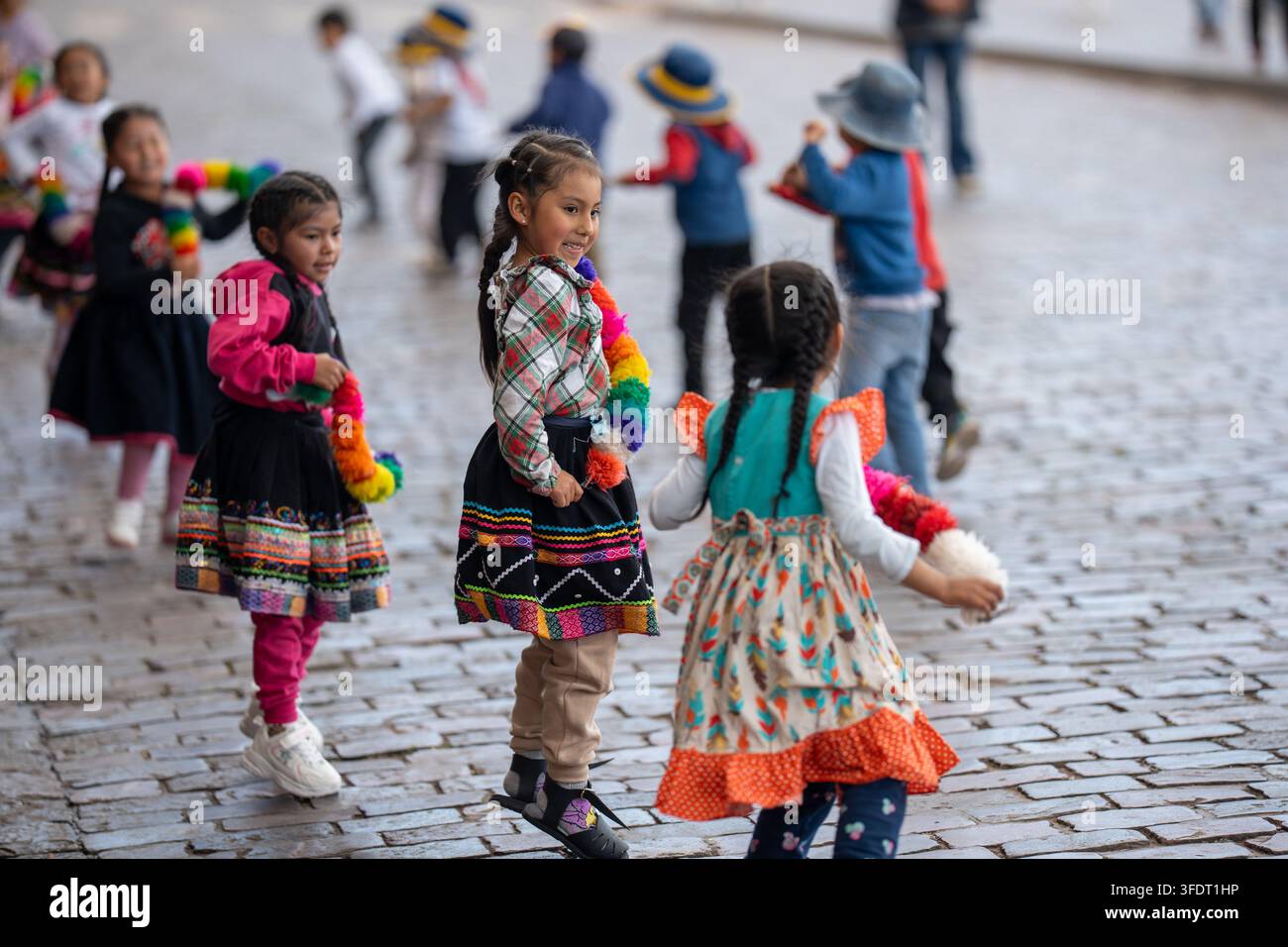 Un gruppo di bambini sorridenti in colorati costumi tradizionali andini danza e gioca su strade di ciottoli durante un vivace festival a Cusco, Perù, cap Foto Stock