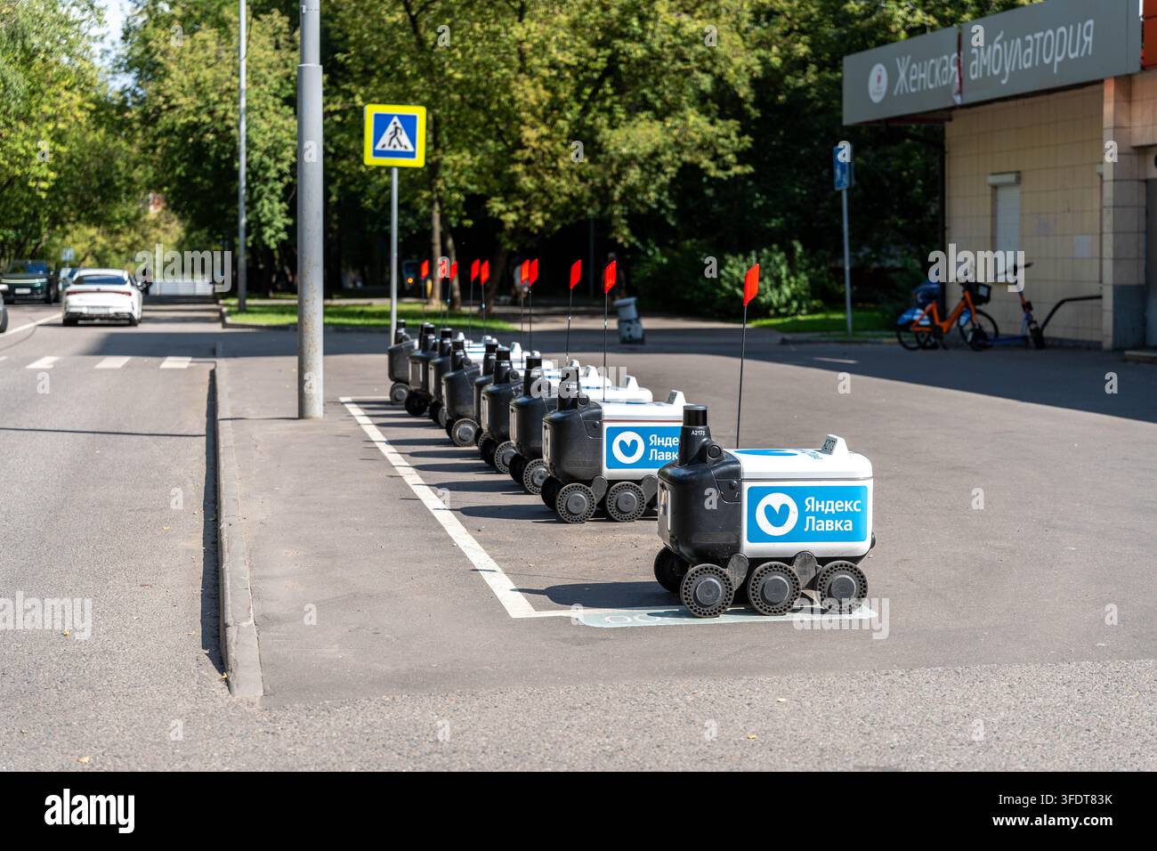 I robot di consegna Yandex Lavka si allineano sul marciapiede accanto al passaggio pedonale su una strada cittadina soleggiata. Piccole unità autonome sono disposte in fila ordinata vicino a un edificio clinico e agli alberi Foto Stock