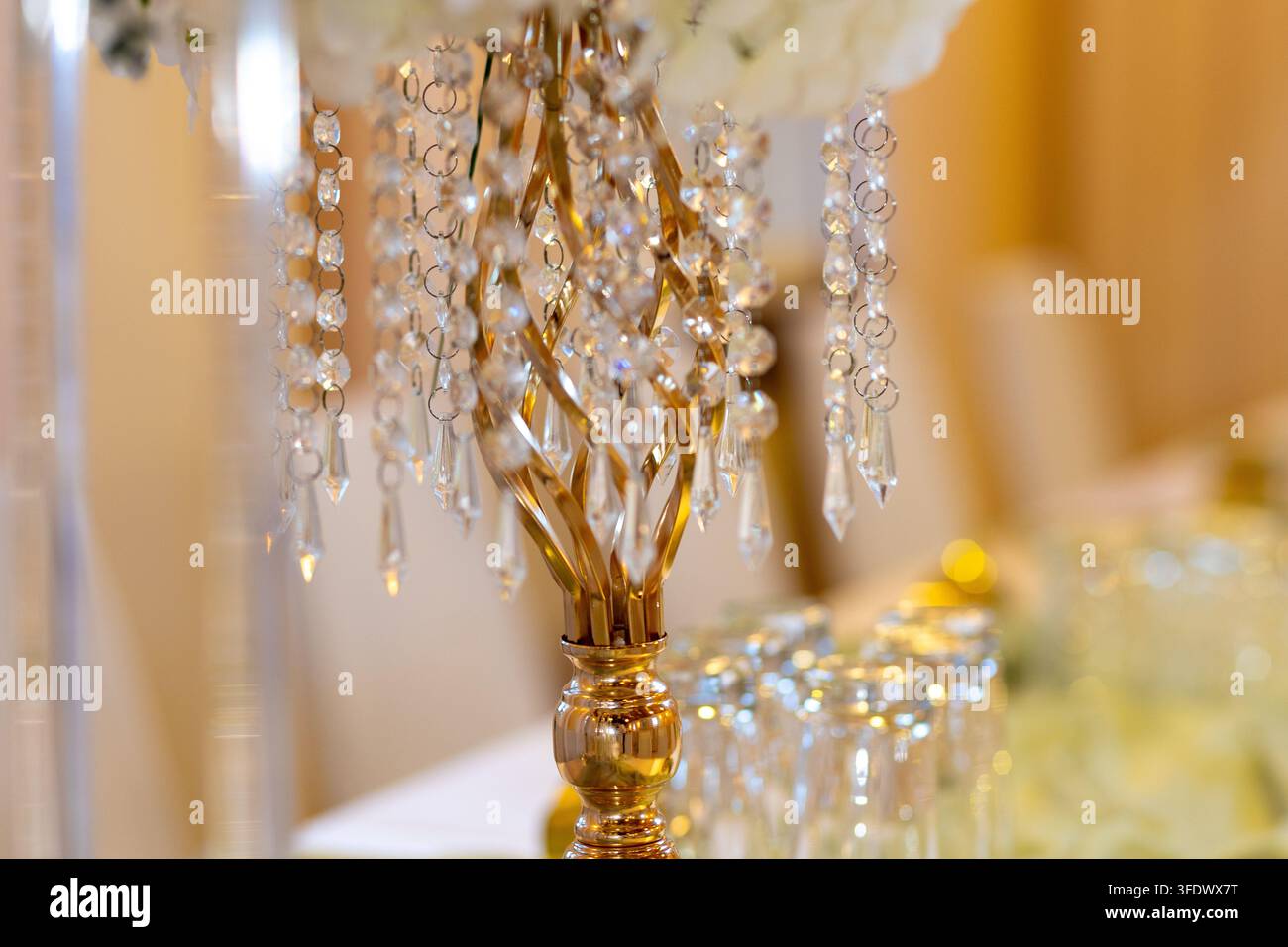 Un centrotavola in oro e cristallo è posato su un tavolo circondato da fiori luminosi. In fondo, durante l'evento, sono disposti bicchieri in fila ordinata. Foto Stock